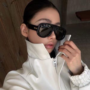 May include: Oversized black sunglasses with a thick frame and dark tinted lenses paired with a white high-collar jacket featuring a silver metal zipper pull. The jacket has a structured collar and gathered cuffs, providing a clean and neutral aesthetic.
