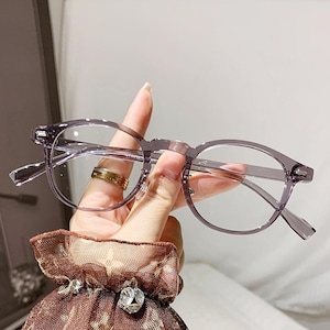 May include: A pair of eyeglasses with a translucent, light purple, rounded plastic frame. The glasses feature clear lenses and thin, matching purple temples. The frame has a smooth, glossy finish and a classic, rounded shape with a bridge.