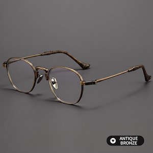May include: Antique bronze spectacles featuring a rounded frame with a textured finish on the rims and bridge. The thin metal temples transition into dark, mottled brown acetate tips. The frame includes adjustable nose pads and clear lenses.