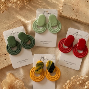 May include: Four pairs of Nara handcrafted polymer clay earrings on white display cards. Each pair features a smooth oval top piece and a textured circular bottom piece. Colours include light green, dark green, bright red, and yellow with black swirls.