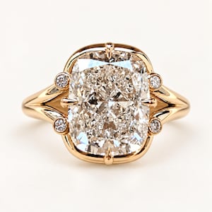 May include: A rose gold ring featuring a large, cushion-cut centre diamond held by four prongs. The setting includes two small, round-cut accent diamonds on each side, integrated into the smooth, polished band that tapers towards the back.