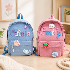 May include: Two corduroy backpacks, one blue and one pink, featuring embroidered names Olivia and Emily. The blue bag has a small flower, rabbit, and elephant patch. The pink bag features a cat face, strawberry, and paw print patch.