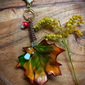 Op de afbeelding: Een sleutelhanger met een groot, meerkleurig esdoornblad in groen-, oranje- en bruintinten. Het blad is bevestigd aan een bruin gevlochten koord met een zilverkleurige metalen kraal, een kleine rood-witte paddenstoelbedel en een grotere oranje paddenstoel.