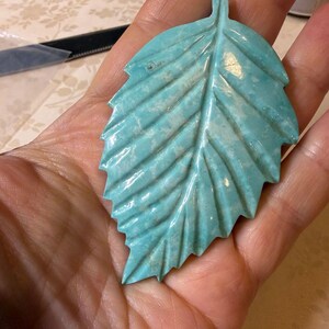 May include: A carved turquoise-coloured leaf pendant with a smooth, glossy finish and visible veining. The leaf features a central stem and detailed serrated edges, showcasing a marbled pattern of light and dark blue-green tones throughout.