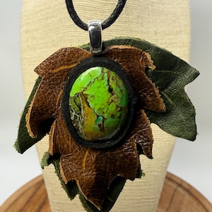 May include: A pendant necklace featuring a polished oval green and brown stone cabochon set in a black frame, layered over brown and dark green leather leaf-shaped cutouts, suspended from a black cord with a silver-toned metal bail.