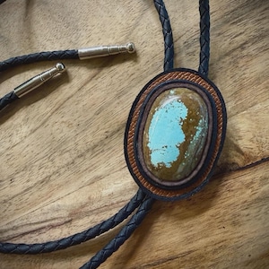 May include: A bolo tie featuring a large oval turquoise and brown stone centerpiece set within layered black and tan leather borders. The cord is made of braided black material and finished with polished silver-toned metal tips at the ends.
