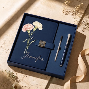 Könnte beinhalten: Ein marineblaues Geschenkset bestehend aus einem Notizbuch und einem passenden Stift. Der Einband des Notizbuchs zeigt eine Aquarellillustration von zwei Nelken, einen goldenen geometrischen Verschluss und den Namen Jennifer in weißer Schreibschrift.