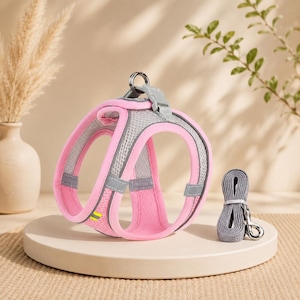 May include: A pink and grey mesh pet harness featuring reflective strips, a metal D-ring for lead attachment, and a plastic buckle closure, accompanied by a matching grey lead with a metal swivel snap hook for secure pet walking.