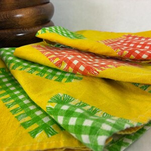 May include: A set of folded yellow fabric napkins featuring a printed pattern of green and red gingham check squares. The napkins have a textured appearance with visible fabric weave and are stacked together in a layered arrangement.