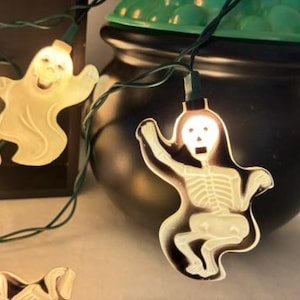 May include: A string of Halloween decorative lights featuring translucent white ghost and skeleton figures. The lights are connected by a dark green wire, with each figure illuminated from within. A black plastic cauldron with a green lid sits behind.