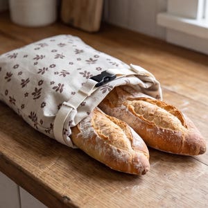 May include: A beige fabric bread bag featuring a repeating brown floral and leaf pattern. The bag is secured with a white adjustable strap and a black plastic side-release buckle, containing two golden-brown crusty baguettes inside.