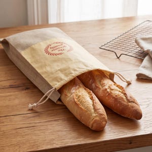 May include: A rectangular beige linen bread bag with a drawstring closure, featuring a yellow panel printed with a red laurel wreath and the text The genuine quality. Two golden-brown crusty baguettes are partially inside the fabric pouch.