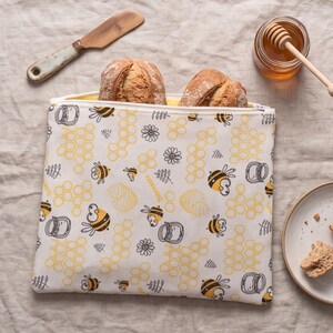 May include: A square fabric food storage bag featuring a repeating pattern of yellow honeycombs, black and yellow cartoon bees, honey jars, and small flowers on a white background, containing two crusty bread loaves with a zipper closure.