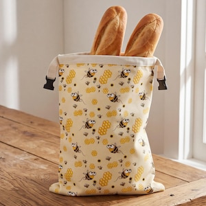 Könnte beinhalten: Ein cremefarbener Stoffbeutel für Brot mit einem wiederkehrenden Muster aus Cartoon-Bienen, gelben Waben und kleinen Sonnenblumen. Der Beutel hat eine obere Öffnung mit zwei schwarzen Kunststoff-Steckschnallen und enthält zwei goldbraune Baguettes.