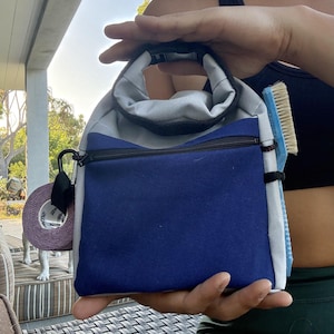 Könnte beinhalten: Eine graue und blaue Segeltuchtasche mit einem Tragegriff, einer Reißverschlusstasche auf der Vorderseite und einer seitlichen Befestigung. Eine Rolle violettes Sporttape ist links befestigt und eine hellblaue Bürste steckt rechts.