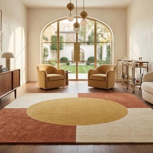 May include: A rectangular area rug featuring a large central yellow circle set against a geometric background of cream and terracotta-coloured quadrants. The rug is positioned on a dark wood floor in a room with a cream sofa and two yellow armchairs.