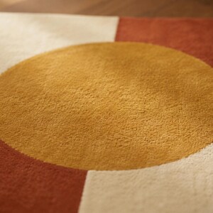 May include: A square rug featuring a large, centred mustard yellow circle set against a background of alternating cream and terracotta red geometric blocks. The rug has a dense, textured pile surface with clean, sharp edges between the colours.