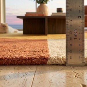 May include: A textured area rug featuring large colour-blocked sections in rust orange, cream, and yellow. A metal ruler is positioned vertically against the edge of the rug, showing a pile height of approximately 1.3 centimetres from the floor surface.