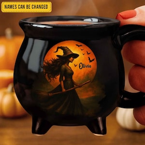 May include: A black cauldron-shaped mug featuring a graphic of a witch in a pointed hat holding a broomstick against a large orange full moon with flying bats. The name Olivia is printed in black script. A yellow box states NAMES CAN BE CHANGED.