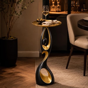 Può includere: Un tavolino scultoreo alto con esterno nero opaco e finitura interna color oro metallizzato. La base ha una forma organica fluida con un'apertura circolare e sostiene un piano d'appoggio rotondo e piatto di colore oro.