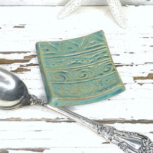 May include: A silver-toned metal spoon with an ornate handle and a stamped inscription in the bowl, paired with a small, square, teal-colored ceramic spoon rest featuring embossed horizontal lines and swirl patterns on its concave surface.