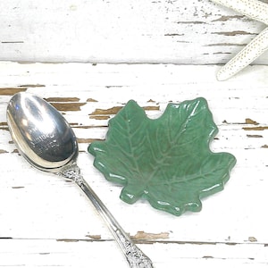 May include: A silver-toned metal spoon with an ornate handle design positioned next to a small, green, leaf-shaped ceramic dish. The spoon has a polished, reflective bowl, and the dish features embossed vein details across its surface.