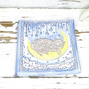 May include: Square ceramic dish with a hand-painted design. The image features a gray cat sleeping on a yellow crescent moon, surrounded by stars and clouds, set against a blue background. The dish has a blue border.