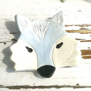 May include: A ceramic dish shaped like a fox head, featuring a light blue forehead and ears, cream-colored cheeks, and a black nose. The fox has two small black eyes and textured fur details carved into the top of the head, creating a flat surface.