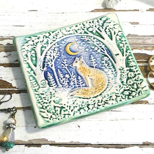 May include: A square ceramic trinket dish featuring a hand-painted scene of a fox sitting in a forest under a crescent moon. The dish has a raised green border and white textured trees. Beside it are two drop earrings and three gold-tone rings.
