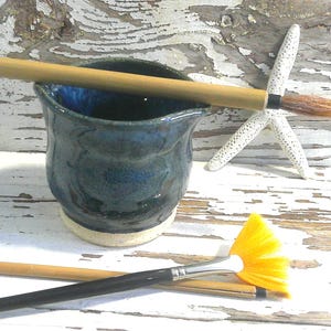 May include: A small, dark blue ceramic pitcher with a glossy finish and a light-colored base. The pitcher is accompanied by two paintbrushes with wooden handles and a white starfish with a textured surface.