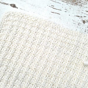 May include: Cream-colored knitted blanket with a textured, diagonal pattern. A matching starfish is placed on the corner of the blanket. The blanket is on a distressed white wooden surface.