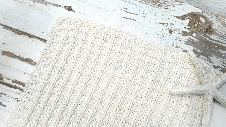 May include: Cream-colored knitted blanket with a textured, diagonal pattern. A matching starfish is placed on the corner of the blanket. The blanket is on a distressed white wooden surface.