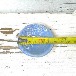 Peut inclure: Un porte-savon en c&eacute;ramique bleu clair avec un motif floral textur&eacute;. Le diam&egrave;tre du plat est d'environ 7,6 cm. Un ruban &agrave; mesurer jaune est plac&eacute; sur le plat, et une &eacute;toile de mer blanche est en arri&egrave;re-plan.