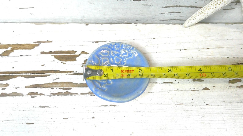 Peut inclure: Un porte-savon en c&eacute;ramique bleu clair avec un motif floral textur&eacute;. Le diam&egrave;tre du plat est d'environ 7,6 cm. Un ruban &agrave; mesurer jaune est plac&eacute; sur le plat, et une &eacute;toile de mer blanche est en arri&egrave;re-plan.