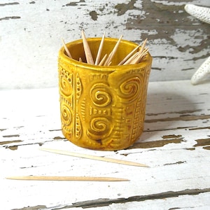 May include: A mustard-yellow ceramic toothpick holder with a carved spiral design. The holder is filled with wooden toothpicks. Two toothpicks are lying on the white, distressed wooden surface. A white starfish is in the background.