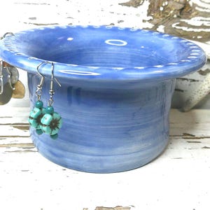 May include: A light blue ceramic container with a decorative rim, holding a pair of turquoise and silver earrings. Additional earrings with circular, metallic accents hang on the side. A white starfish rests in the background.
