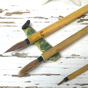 May include: Three calligraphy brushes with bamboo handles and brown bristles rest on a green and yellow ceramic brush rest. The brushes are arranged on a distressed white wooden surface.