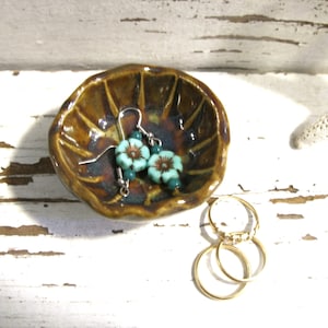 May include: A small, handcrafted ceramic dish with a brown glaze, holding a pair of turquoise flower earrings and three gold rings. The earrings feature turquoise flower beads and dark green beads. The dish sits on a white, distressed wooden surface.