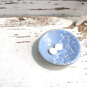 Peut inclure: Un petit plat en c&eacute;ramique bleu clair avec un motif textur&eacute;. Deux objets blancs et ronds reposent au centre du plat. Le plat est pos&eacute; sur une surface en bois blanc vieilli.