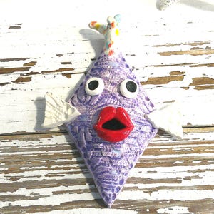 May include: A whimsical, diamond-shaped ceramic fish with a textured, lavender body. It features large, white-rimmed eyes, a bright red open mouth, and white fins. The fish has a colorful, dotted fin and is a decorative item.
