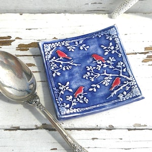 May include: A square ceramic trinket dish with a deep blue glaze featuring a raised white tree branch pattern and four small red birds perched on the branches, positioned next to a polished silver metal spoon with an ornate handle design.