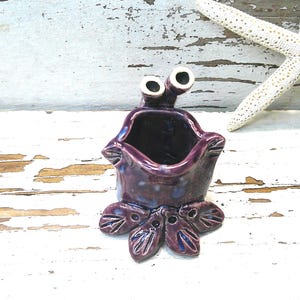 May include: A whimsical, handmade ceramic planter in shades of purple and blue. The planter has a unique, stylized design with large, round eyes and a petal-like base. A white starfish is in the background.