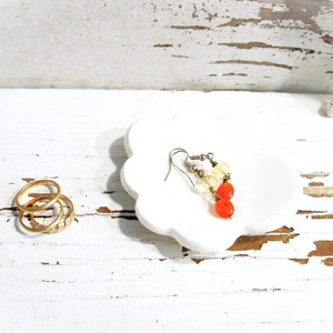May include: A white, cloud-shaped trinket dish holds a pair of earrings with orange and yellow beads. Three gold rings are to the left of the dish. The background is a white, distressed wooden surface with a starfish.