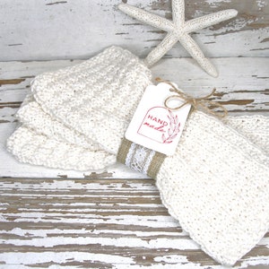 May include: Off-white, hand-knitted washcloths are stacked and tied with a burlap and lace ribbon. A tag with the words "HAND made" is attached. A white starfish is in the background. The washcloths are on a distressed white wooden surface.