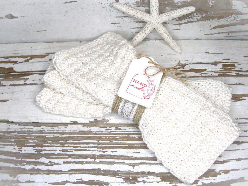 May include: Off-white, hand-knitted washcloths are stacked and tied with a burlap and lace ribbon. A tag with the words "HAND made" is attached. A white starfish is in the background. The washcloths are on a distressed white wooden surface.
