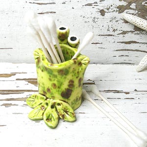 May include: A whimsical, handmade ceramic frog-shaped container, speckled with green and brown, holds white cotton swabs. A starfish rests nearby on a distressed white wooden surface. The frog has leaf-shaped feet.