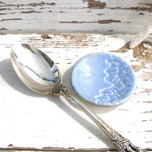 Peut inclure: Une cuill&egrave;re de service en argent avec des d&eacute;tails orn&eacute;s est pos&eacute;e &agrave; c&ocirc;t&eacute; d'une petite coupelle ronde en c&eacute;ramique bleu clair avec un motif textur&eacute;. La cuill&egrave;re et le plat sont sur une surface en bois blanc vieilli.