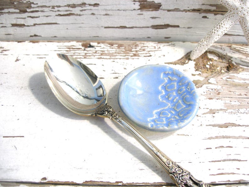 Peut inclure: Une cuill&egrave;re de service en argent avec des d&eacute;tails orn&eacute;s est pos&eacute;e &agrave; c&ocirc;t&eacute; d'une petite coupelle ronde en c&eacute;ramique bleu clair avec un motif textur&eacute;. La cuill&egrave;re et le plat sont sur une surface en bois blanc vieilli.