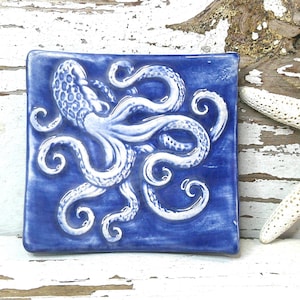 May include: A square ceramic dish featuring a raised white octopus relief design against a deep blue glazed background. The octopus has detailed tentacles and a textured head, creating a three-dimensional effect on the glossy surface of the tray.
