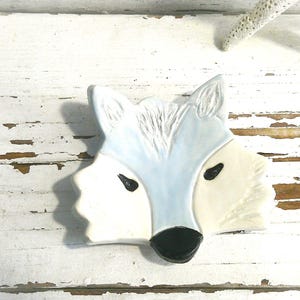 May include: A ceramic fox head sculpture in white, light blue, and cream. The fox has black eyes and a black nose. The sculpture is placed on a distressed white wooden surface. A white starfish is in the background.